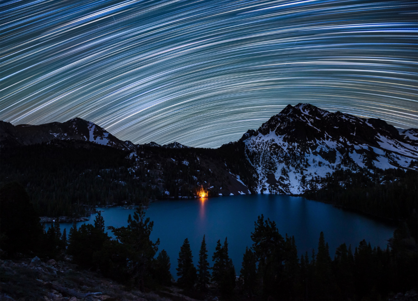 Star trails illuminate the night sky over a campfire-lit Green Lake in the Hoover Wilderness of California. S