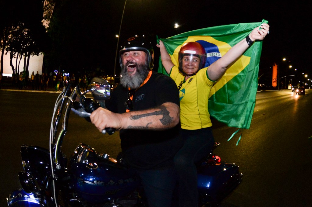 Two people riding a motorised scooter holding a Brazilian flag.